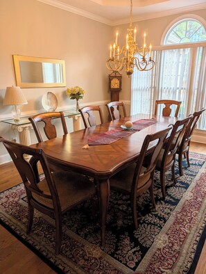 A dining room with a large wooden table, six chairs, a chandelier, a clock, and a window with blinds.