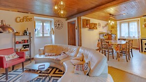 Een gezellige woonkamer met een gebogen witte bank, een rode fauteuil, een muur met koffie thema, en een eethoek met een tafel en stoelen.