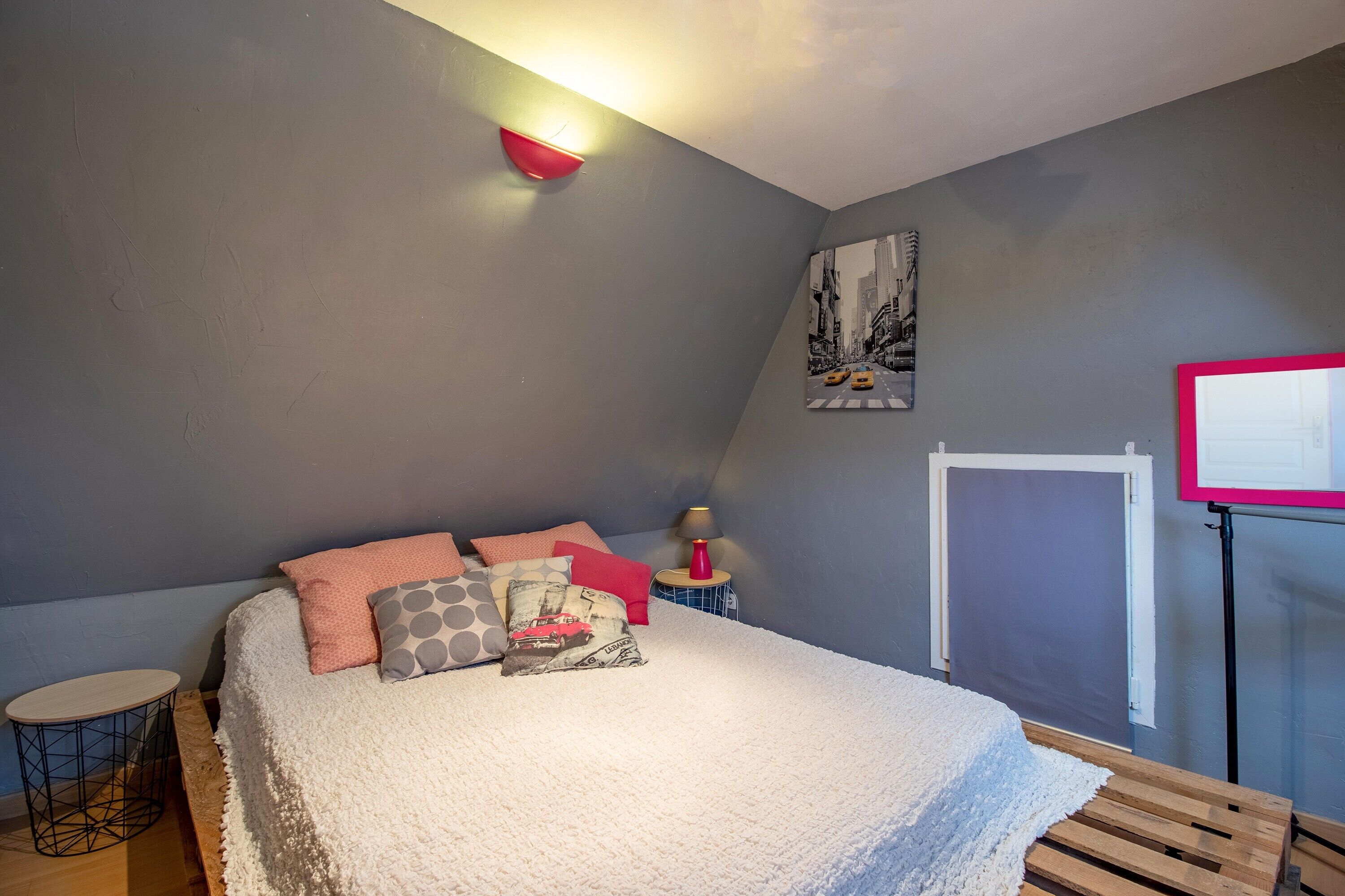 Ein Schlafzimmer mit Bett, Kissen, einem Nachttisch mit Lampe, einem an der Wand befestigten Regal und einem Fenster mit Jalousie.