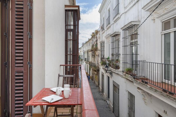 A narrow urban street with a red table and chairs on a balcony, white buildings with balconies, and a clear sky.