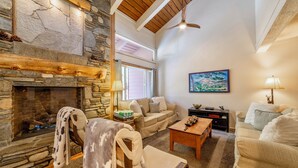A cozy living room with a stone fireplace, wooden beams, a ceiling fan, and a painting on the wall. - Updated Ski-Back Condo w/ Garage at Canyon Lodge (Mammoth Lakes)