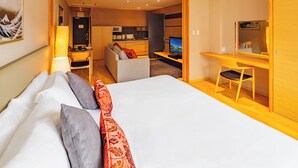 A modern hotel room with a large bed, a desk, a chair, a TV, and a kitchenette in the background.