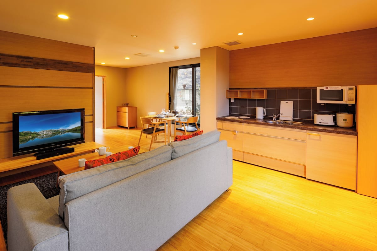 Een moderne keuken met houten kasten, een eethoek met een tafel en stoelen, een grijze bank en een televisie.