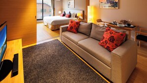 A hotel room with a bed, a sofa, a TV, a dining table with chairs, and a painting on the wall.
