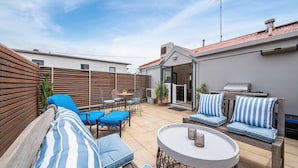 A patio area with a wooden bench, blue striped cushions, a round table with two candles, and a grill.
