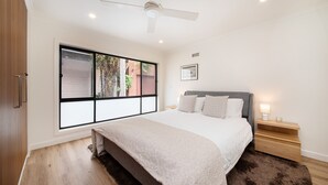 A bedroom with a large window, a bed, a bedside table with a lamp, and a ceiling fan.