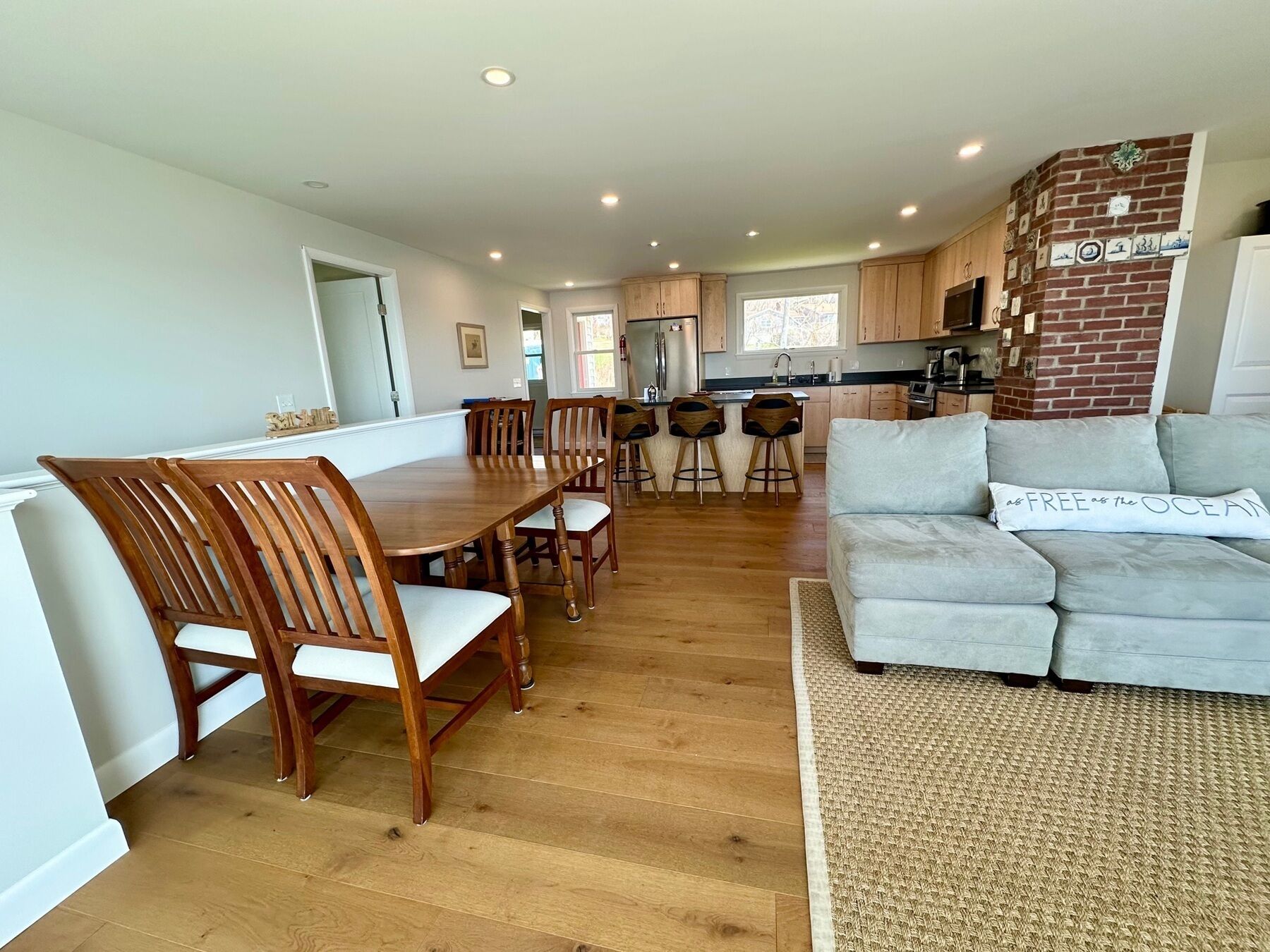 A modern kitchen with wooden cabinets, a dining area with a wooden table and chairs, and a living room with a grey sofa.