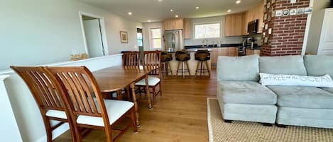 A modern kitchen with wooden cabinets, a dining area with a wooden table and chairs, and a living room with a grey sofa.