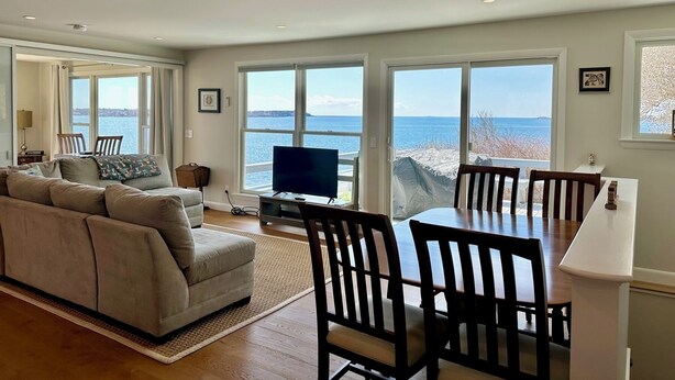 A spacious living room with a large sofa, a dining table with chairs, a flat-screen TV, and large windows offering a view of the sea. - Stunning Ocean Views, newly renovated cottage-close to preserves, trails, and Harpswell Beaches (Harpswell)
