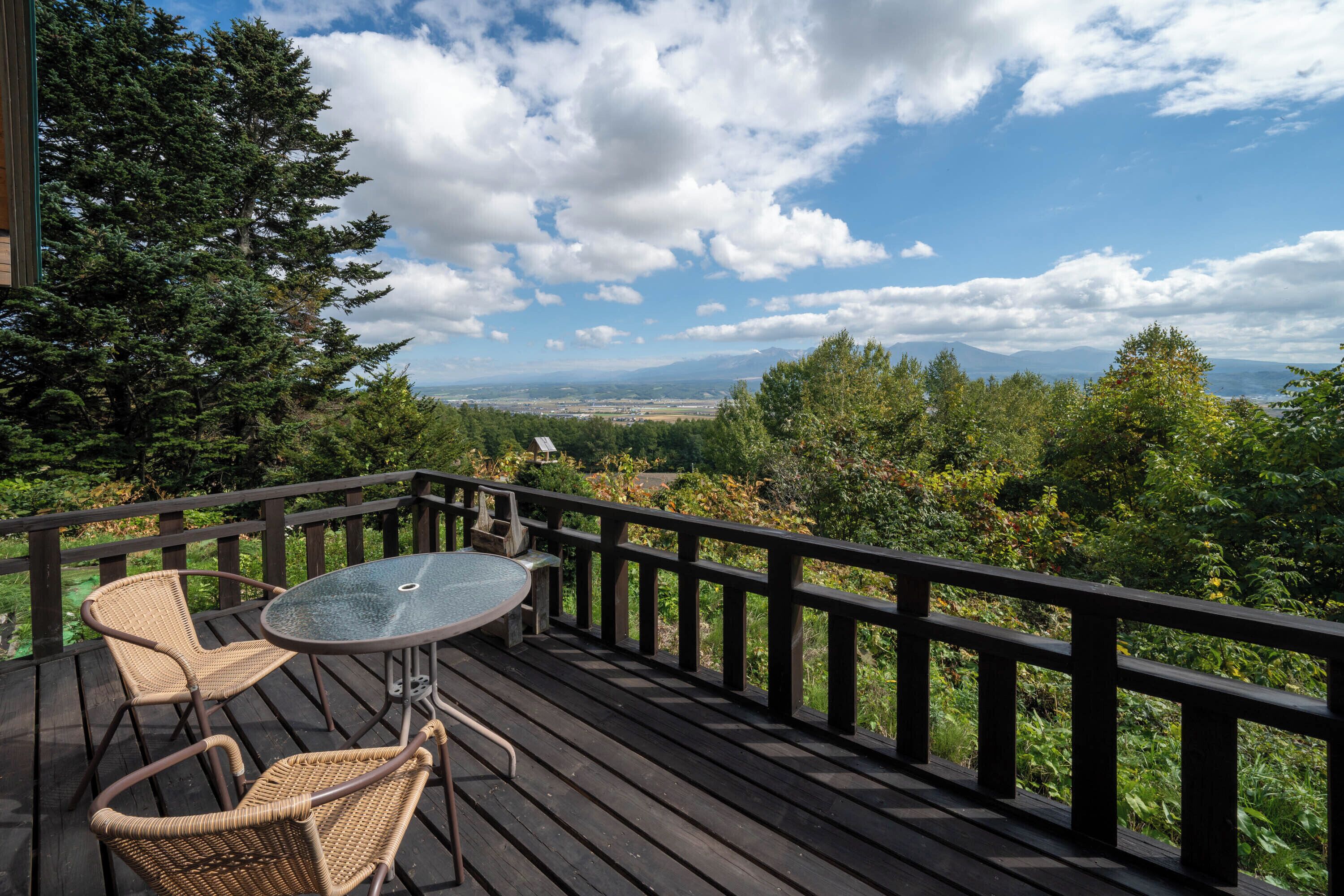 A wooden deck with a glass table and wicker chairs, overlooking a scenic landscape with trees and mountains.