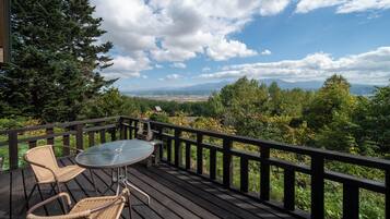A wooden deck with a glass table and wicker chairs, overlooking a scenic landscape with trees and mountains.
