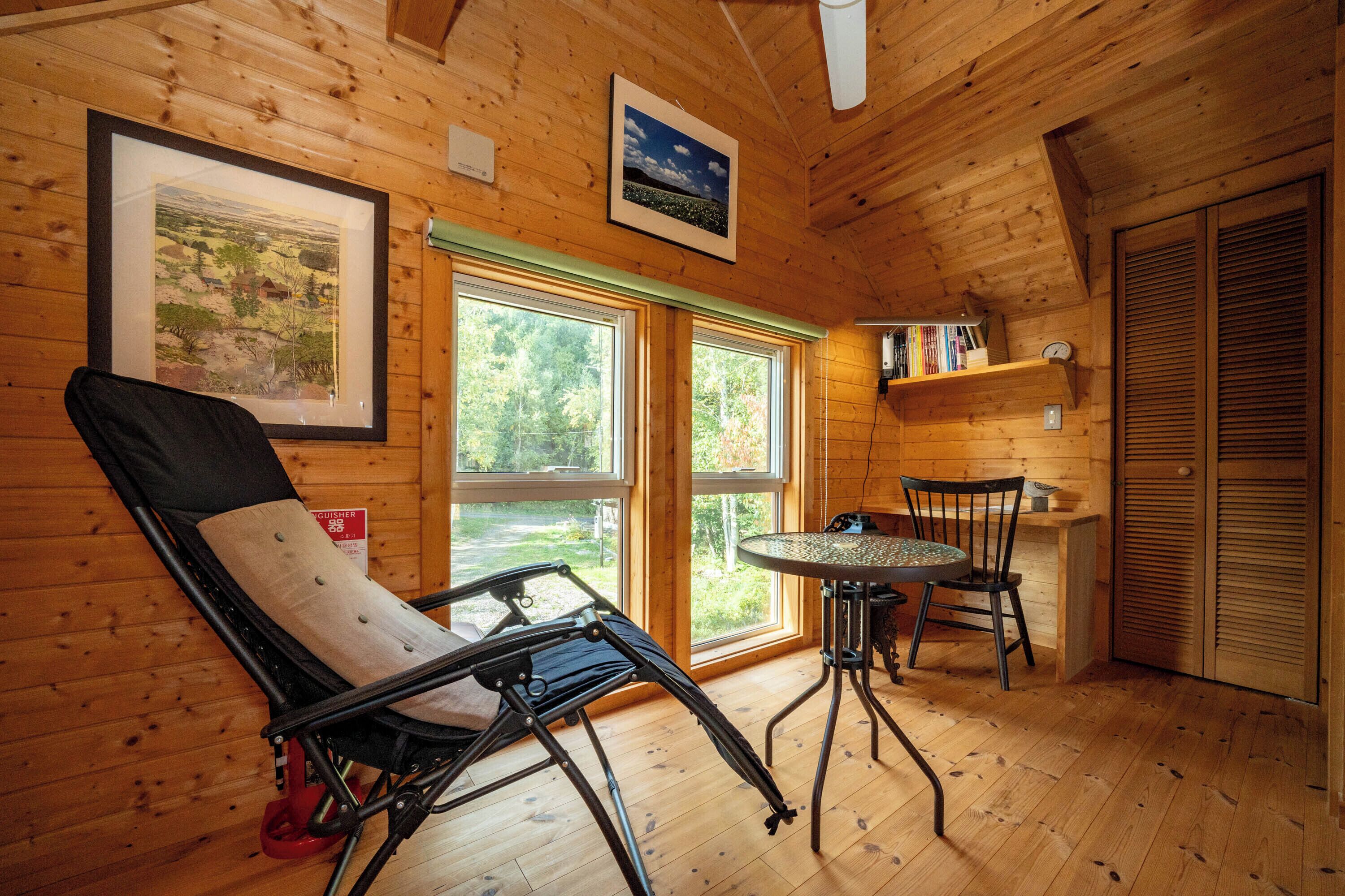 A wooden cabin interior with a reclining chair, a small table, and chairs, large windows, and a door with shutters.