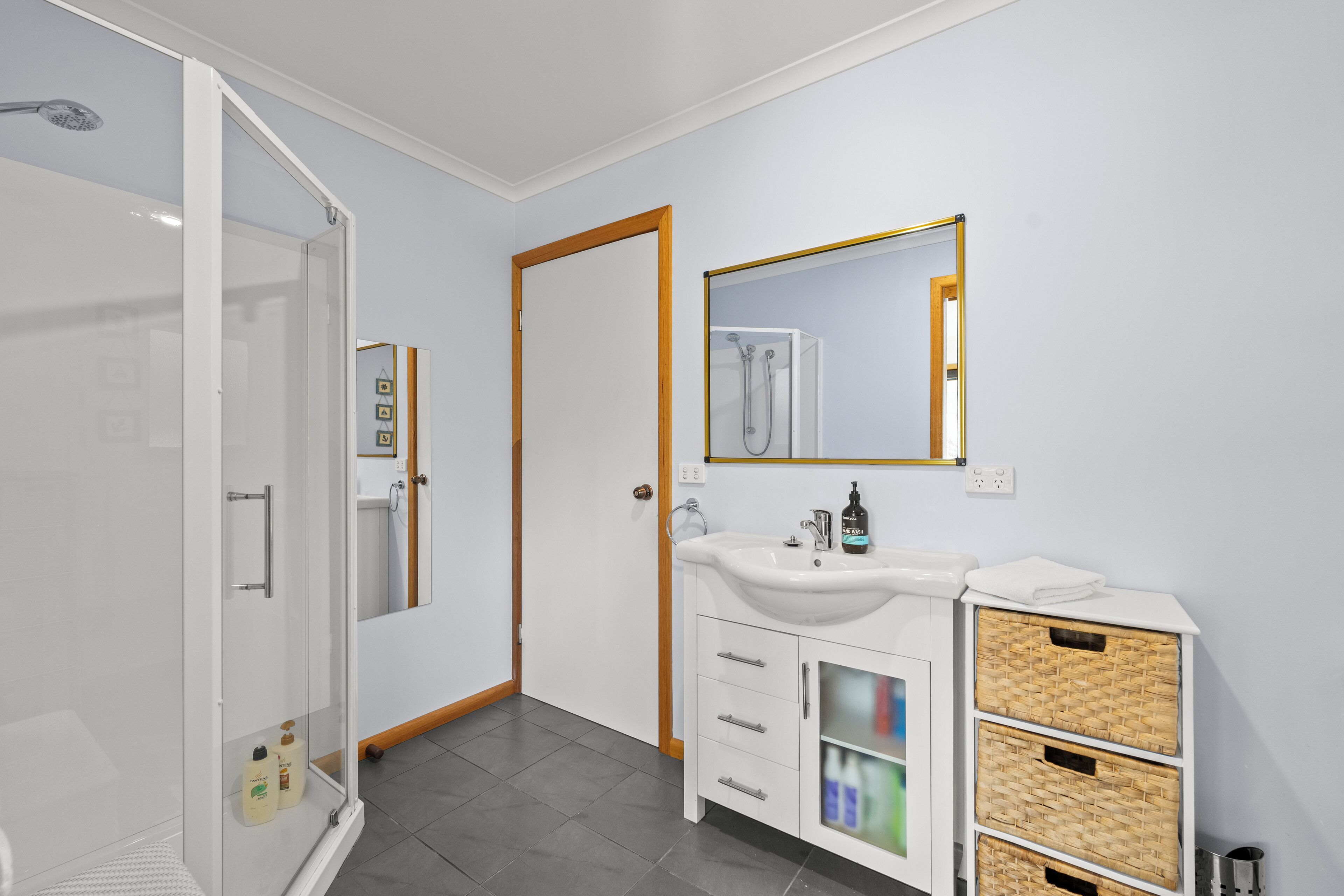 Un baño moderno con cabina de ducha de vidrio, lavamanos blanco con espejo y cestas de mimbre a los lados.