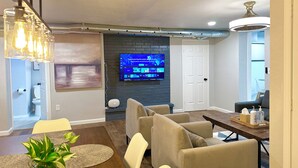 A modern living room with a flat-screen TV mounted on a brick wall, a dining area with a wooden table and beige chairs, and a ceiling fan. - 7BR Comfort Haven For 16 I Next to Tinker I EC (Midwest City)