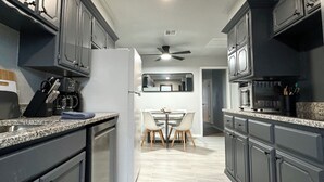 A modern kitchen with dark cabinetry, a refrigerator, a coffee maker, and a dining area with a table and chairs. - 7BR Comfort Haven For 16 I Next to Tinker I EC (Midwest City)