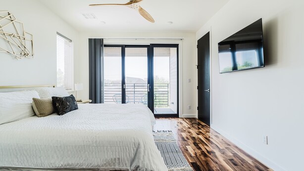 A bedroom with a bed, a ceiling fan, a TV, a large window, and a door leading to a balcony. - Cozy Getaway Perfect for 10 Prime Location NW6 (Oklahoma City)