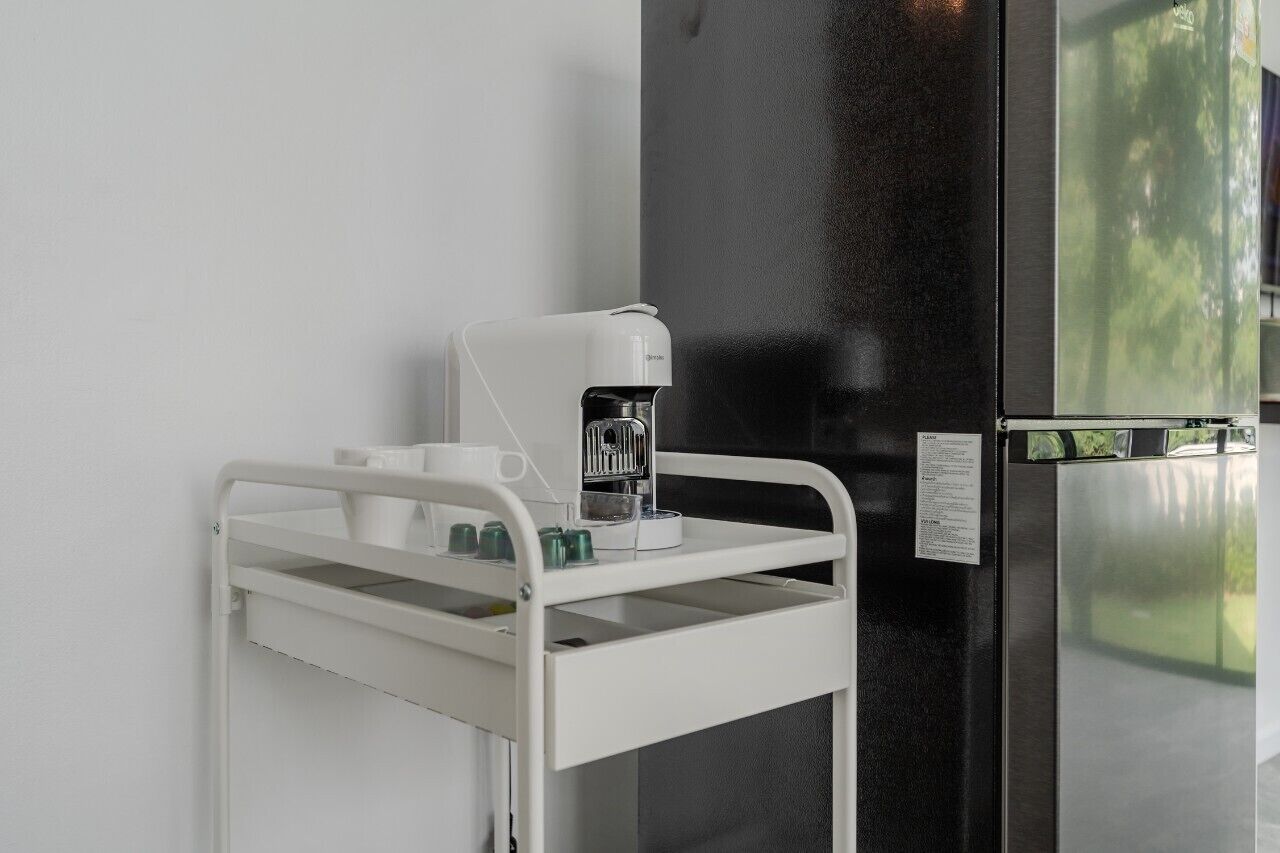 Een witte kar met een koffieapparaat en bekers, geplaatst naast een zwarte koelkast in een keuken.