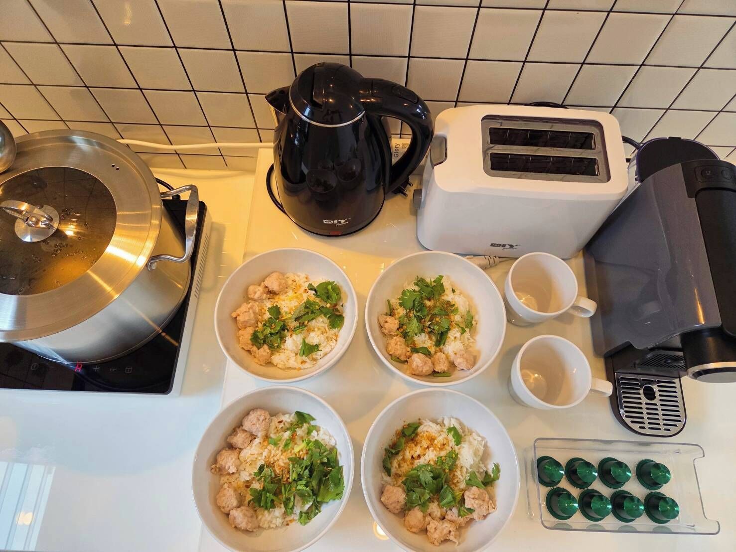 A kitchen counter with four bowls of rice topped with meat and herbs, a kettle, a toaster, and a coffee machine.