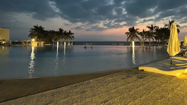 A pool area with palm trees and umbrellas, a beach, and a sunset sky.