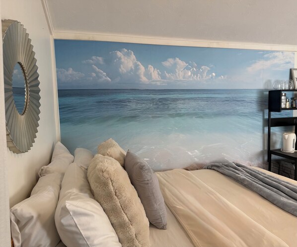 A bedroom with a large ocean view mural, a bed with multiple pillows, a nightstand, and a shelf with items. - Oceanfront Studio :Ocean access, boardwalk, dock, BBQ, Kayaks & crab traps! (Sooke)