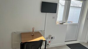 A room with a wooden table, a black chair, a TV mounted on the wall, a window with a view of a garage, and a door with a mat. - Ldn Studio-Free Parking-15 min to Elizabeth Line (Dagenham)