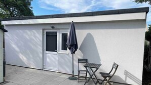 A small, single-story building with a white exterior, a door, and a window. There is a small patio area with a table and two chairs, and a single umbrella. - Ldn Studio-Free Parking-15 min to Elizabeth Line (Dagenham)