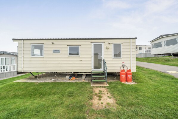 A single-story mobile home with a front door, two windows, and a small porch. There are two propane tanks on the ground beside the home.