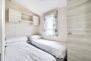 A bedroom with two single beds, a window with floral curtains, and a wooden wardrobe. - PerranporthCaravan/2bedroom/Tv/GasFire (Perranporth)