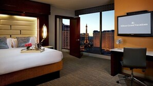 Habitación de hotel con cama, escritorio, silla, pantalla de televisión mostrando 'Hilton Grand Vacations' y vista a un paisaje urbano con un edificio alto.
