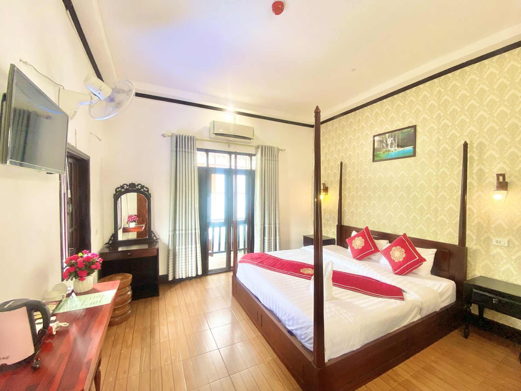 A hotel room with a four-poster bed, a flat-screen TV, an air conditioning unit, a desk with a chair, and a window with curtains.
