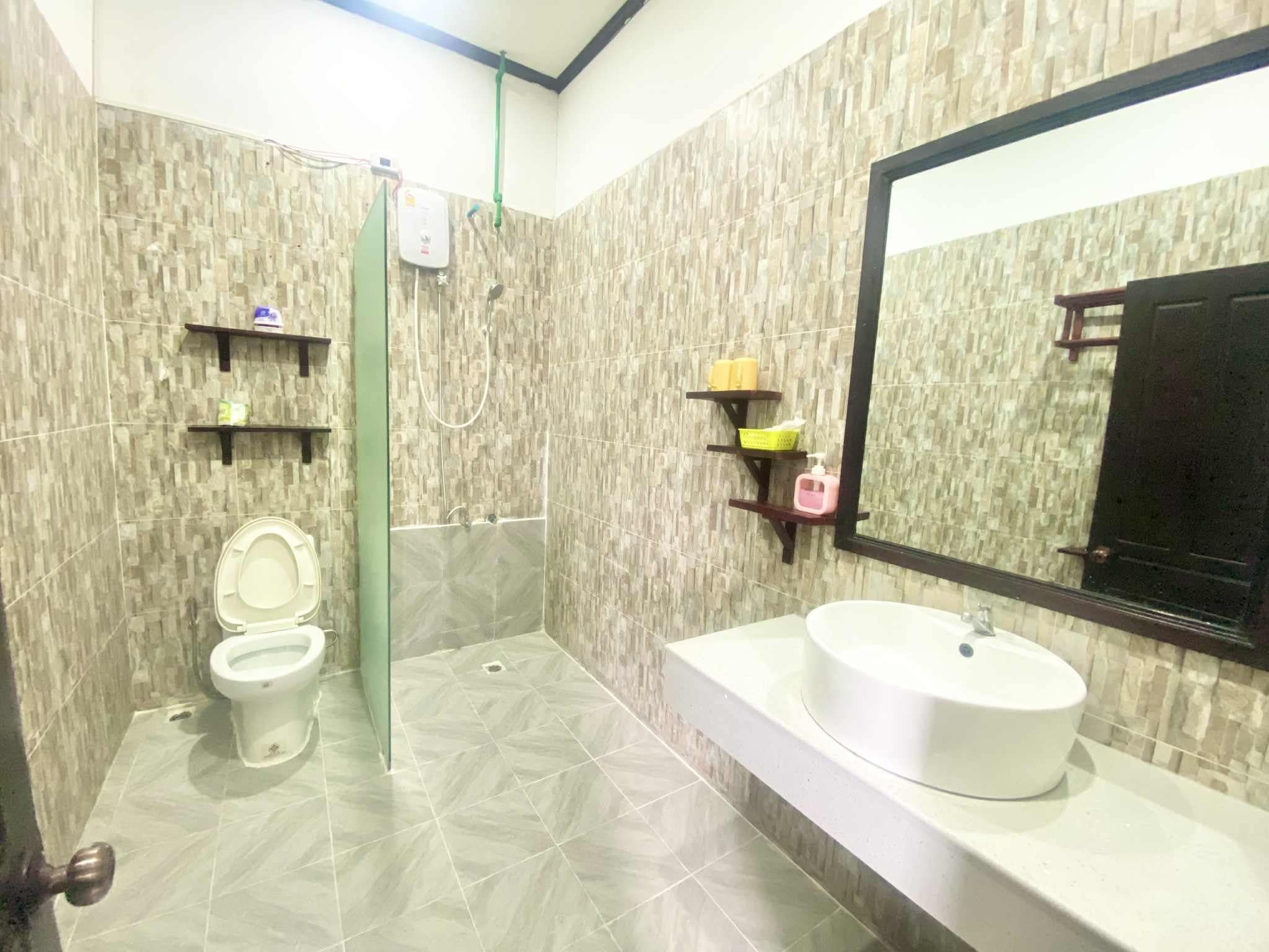 Baño con inodoro, ducha y lavamanos. Hay repisas con artículos de aseo.