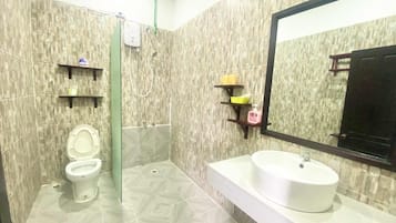 Baño con inodoro, ducha y lavamanos. Hay repisas con artículos de aseo.