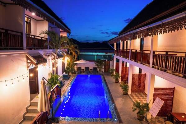 Una piscina con borde de baldosas, rodeada de edificios con balcones y luces colgantes.