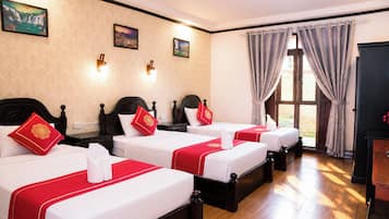 Habitación de hotel con cuatro camas individuales, cada una con cubrecama rojo y blanco, un almohadón blanco y un almohadón rojo con un emblema dorado. Hay luces fijadas a la pared, un cuadro y un ventanal grande con cortinas.