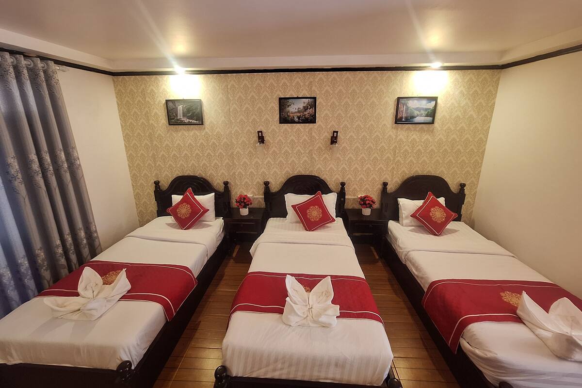 Une chambre d’hôtel avec trois lits simples, chacun recouvert d’une couvre-lit rouge et blanche et agrémenté d’un coussin décoratif. La chambre est dotée d’un plancher en bois, d’un papier peint à motifs et de deux lampes fixées au mur.