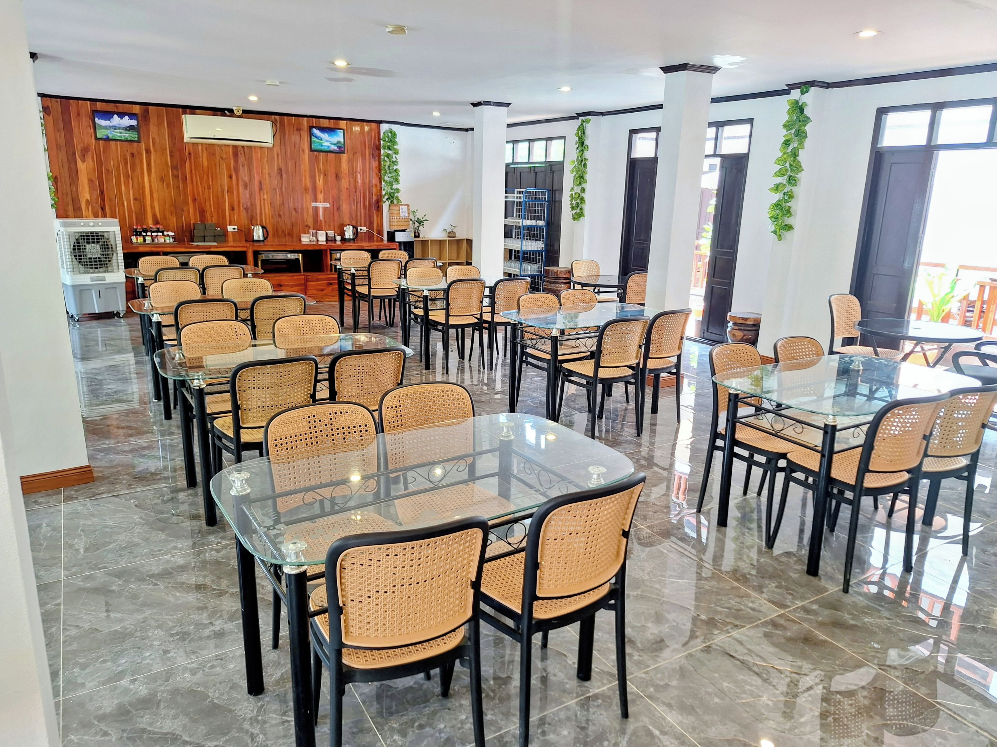 Un vaste espace dédié à la salle à manger, avec des tables en verre et des chaises en osier, des murs lambrissés et un ventilateur de plafond.