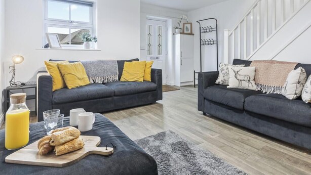 A modern living room with two dark sofas, a wooden coffee table, a staircase, and a kitchen in the background.