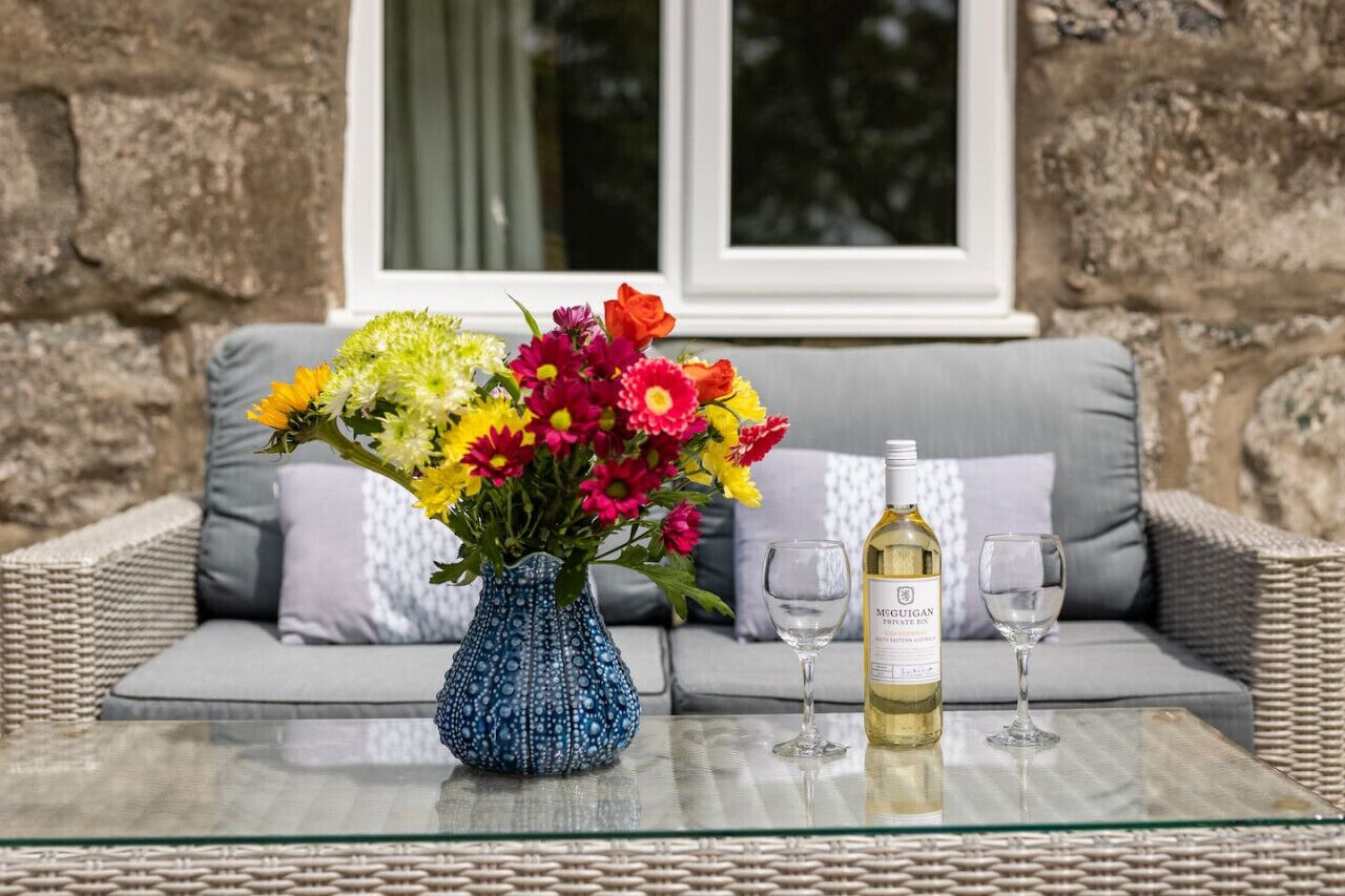 A glass table with a blue vase of flowers, a bottle of wine, and two wine glasses on a wicker sofa with grey cushions.