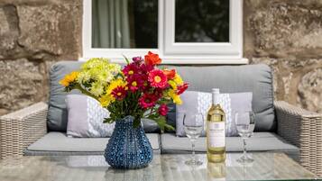 A glass table with a blue vase of flowers, a bottle of wine, and two wine glasses on a wicker sofa with grey cushions.