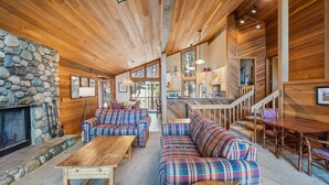 A cozy interior with a stone fireplace, plaid sofas, wooden tables, and a kitchen area with a staircase.