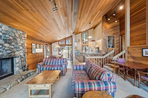 A cozy interior with a stone fireplace, plaid sofas, wooden tables, and a kitchen area with a staircase.