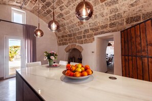 A kitchen with a marble countertop, a bowl of fruit, hanging pendant lights, and a stone wall.