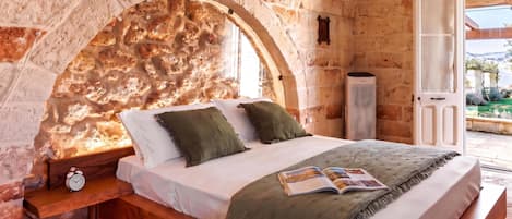 A bedroom with a stone wall, a bed with pillows, a bedside table with a clock, and a view of the outdoors.