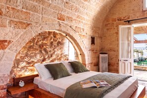 A bedroom with a stone wall, a bed with pillows, a bedside table with a clock, and a view of the outdoors.
