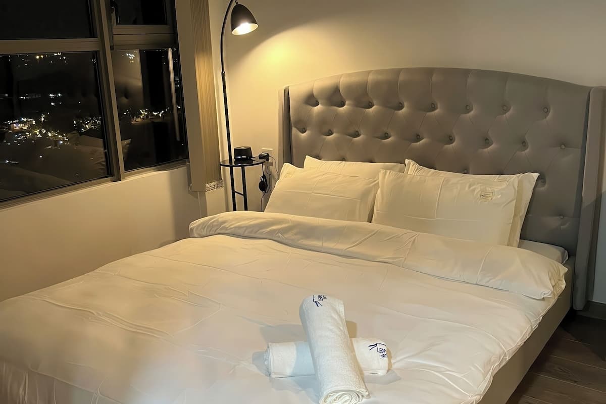 Una cama bien hecha con sábanas blancas y una toalla enrollada, una lámpara de noche y vista a la ciudad de noche a través de una ventana.