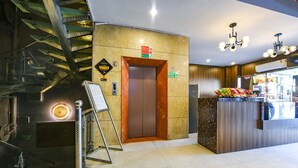 A hotel lobby with a staircase, an elevator, a reception desk, and a fire exit sign.