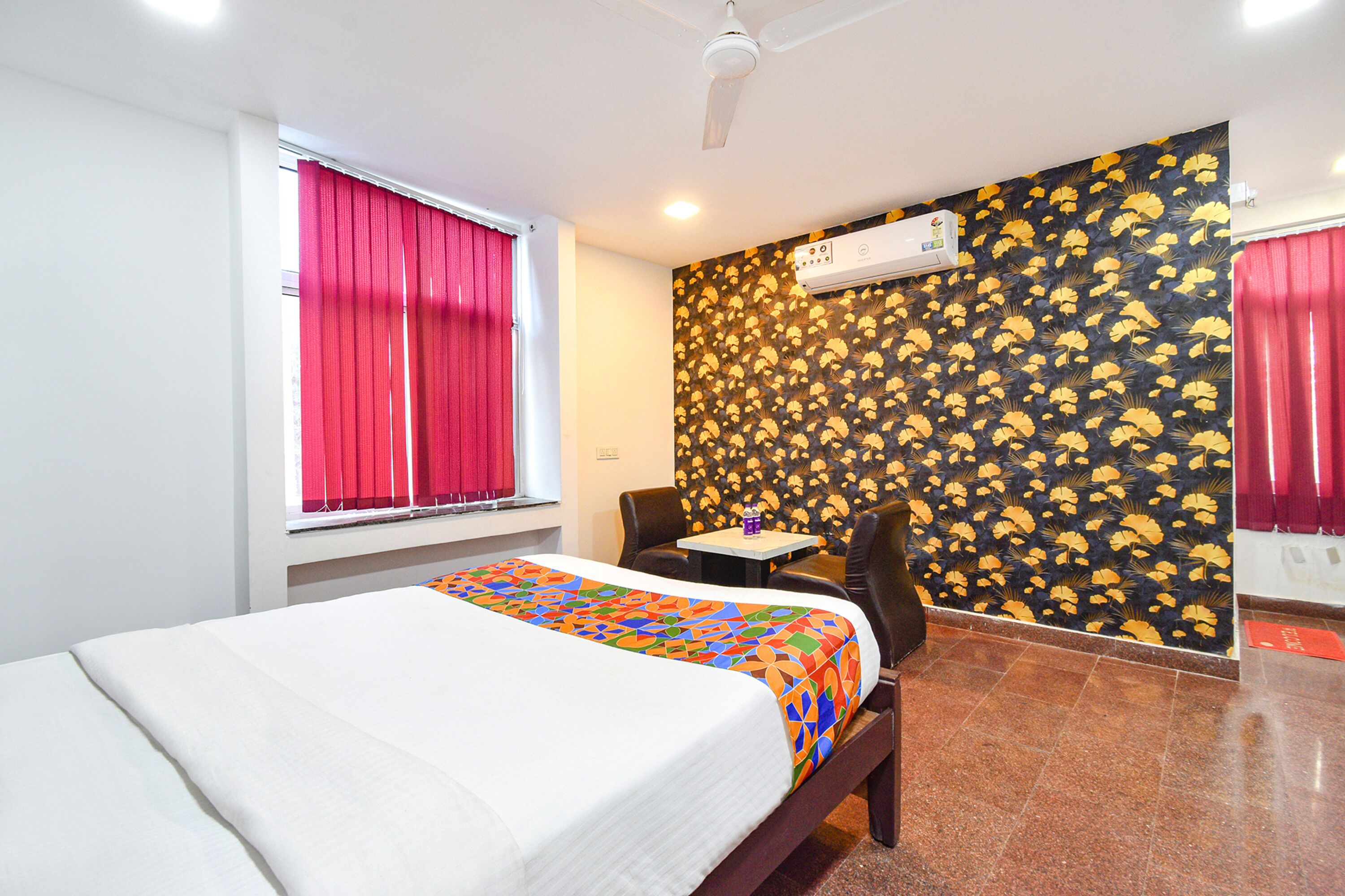 A hotel room with a bed, a small table, a chair, a window with red curtains, and a wall with a floral pattern.