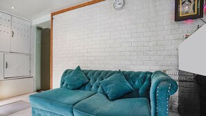 Un canapé recouvert d’un tissu turquoise, adossé à un mur en briques blanches, avec une horloge et un tableau encadré.