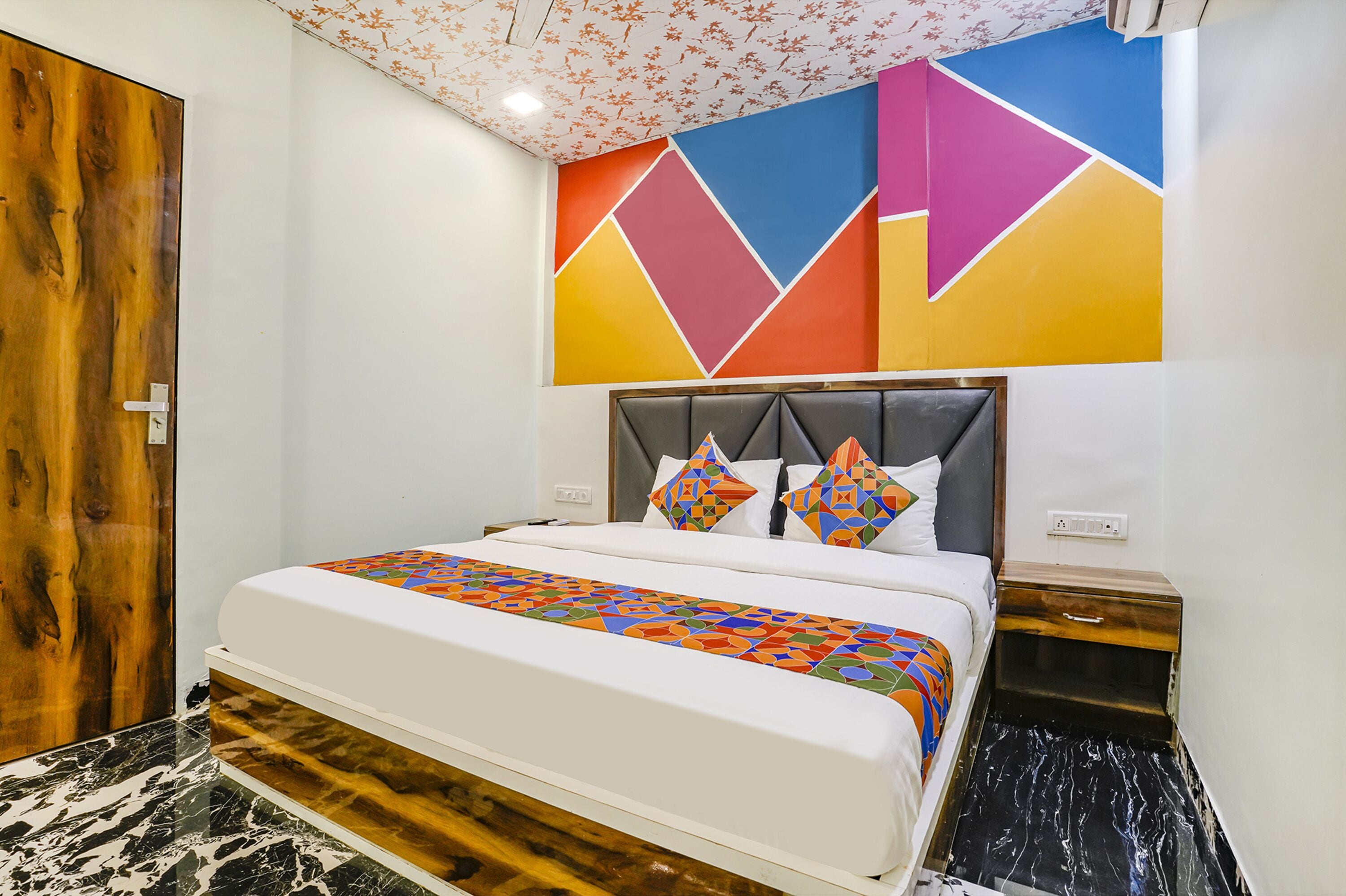 Una habitación de hotel con una cama grande, una puerta de madera, un diseño geométrico colorido en la pared y un piso de mármol.