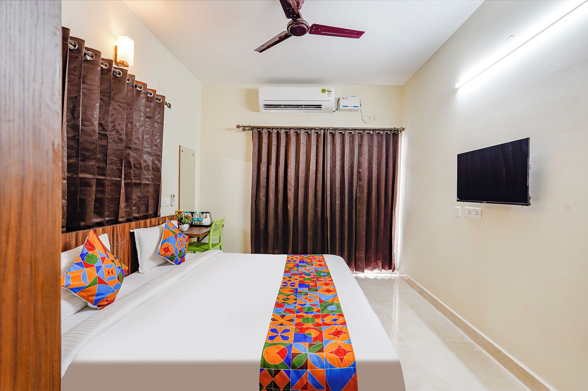 A hotel room with a bed, a desk, a chair, a TV, an air conditioning unit, and a ceiling fan.